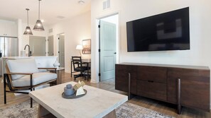 A modern living room with a flat-screen TV mounted on the wall, a dining area with a table and chairs, and a kitchen with a refrigerator and cabinets. - Landing | Spectacular 1BD in Heath Brook (Ocala)