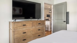 A bedroom with a bed, a wooden dresser with drawers, a television mounted above the dresser, and a door leading to a kitchen area. - Landing | Spectacular 1BD in Heath Brook (Ocala)