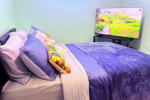 A bed with a purple blanket and multiple pillows, a television displaying a landscape, and a wall socket. - Emerald Calm Retreat | King + Queen | YYC (Calgary)