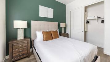 Una cama bien tendida con cabecera capitonada, dos mesitas de noche con lámparas, un armario con espacio para colgar ropa y una puerta que da a un vestidor.
