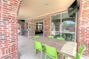 A brick exterior wall with a wooden table and green chairs, a glass door leading inside, and a television mounted on the wall. - Landing | Spectacular Studio, Gym (College Station)
