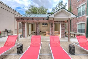 A patio area with red lounge chairs, a barbecue grill, and a covered seating area with tables and chairs. - Landing | Beautiful 1BD, Gym (College Station)