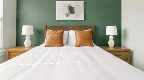 A neatly made bed with white linens, two brown pillows, and a wooden headboard. There are two bedside tables with lamps and a wall-mounted abstract art piece above the bed. - Landing | Beautiful 2BD, Game Room, Gym (Houston)
