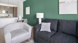 A modern living room with a grey sofa, a grey armchair, a white lamp, and a green accent wall. - Landing | Chic 1BD, Gym, Clubhouse (Houston)