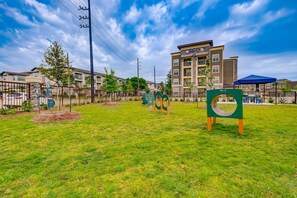 A grassy area with playground equipment, a fence, and a multi-story building in the background. - Landing | Stylish 1BD, Gym, Game Room (Houston)