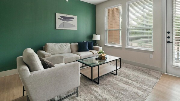 Ein Wohnzimmer mit grüner Wand, einem beigen Sofa, einem Glastisch und einer Tür mit Jalousien.