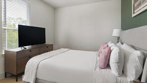 A bedroom with a bed, a TV on a wooden dresser, and a window with blinds. - Landing | Chic 1BD in Deerbrook (Houston)