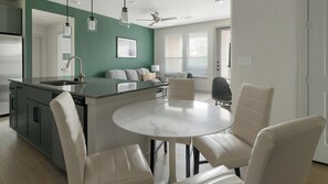 Cozinha moderna com ilha central, sala de jantar com cadeiras e sala de estar com sofá e televisão.