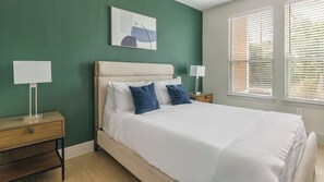 Een slaapkamer met een bed, twee nachtkastjes, een raam met jaloezieën en een aan de muur bevestigd schilderij.