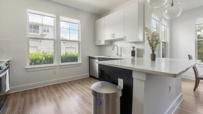 A modern kitchen with white cabinetry, a stainless steel sink, and a large island with a trash can. - Landing | Incredible 2BD, Game Room, Gym (Cape Coral)