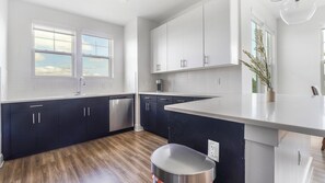A modern kitchen with dark cabinets, a stainless steel dishwasher, and a large island. - Landing | Modern 2BD, Gym, Game Room (Cape Coral)