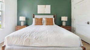 Een net opgemaakt bed met witte lakens en twee bruine kussens, aan weerszijden omringd door houten nachtkastjes met lampen, in een kamer met groene wanden en een witte deur.