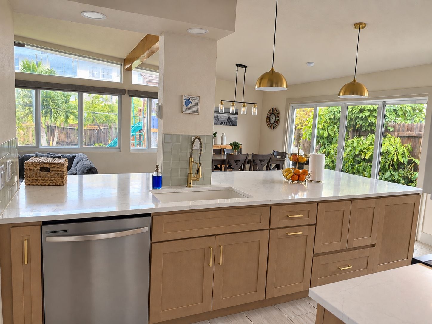 A modern kitchen with a stainless steel dishwasher, wooden cabinets, and a large window overlooking a garden.