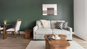 A living room with a grey sofa, a wooden coffee table, a dining table with chairs, and a green accent wall. - Landing | Incredible 1BD, Gym, Game Room (Cape Coral)