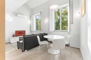 Un salon moderne avec un canapé gris, une table ronde blanche et des chaises, un téléviseur à écran plat et de grandes fenêtres donnant sur la forêt.