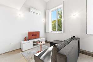 A modern living room with a flat-screen TV, a grey sofa, a white entertainment unit, and a window with a view of trees. - GuestReady-Rou27-G (Porto)
