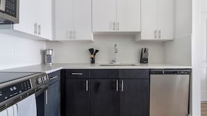 A modern kitchen with white cabinets, a stainless steel dishwasher, and a black stovetop. - Landing | Spectacular 1BD, Gym, Game Room (Cape Coral)