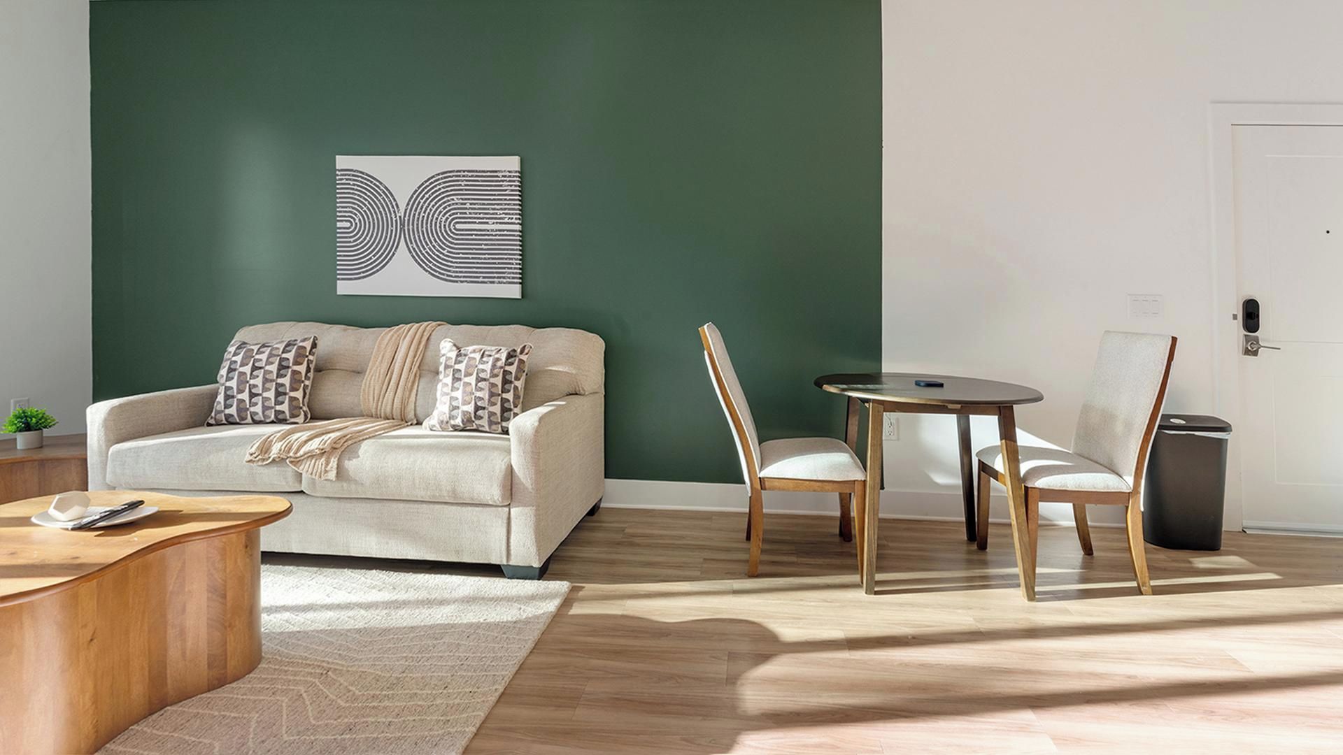 Een moderne woonkamer met een groene accentmuur, een beige bank, gekleurde kussens, een houten salontafel, een ronde eettafel met stoelen en een prullenbak.
