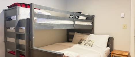Una cama litera con estructura de madera, una cama individual abajo, una mesita de noche de madera y un termostato fijado a la pared.