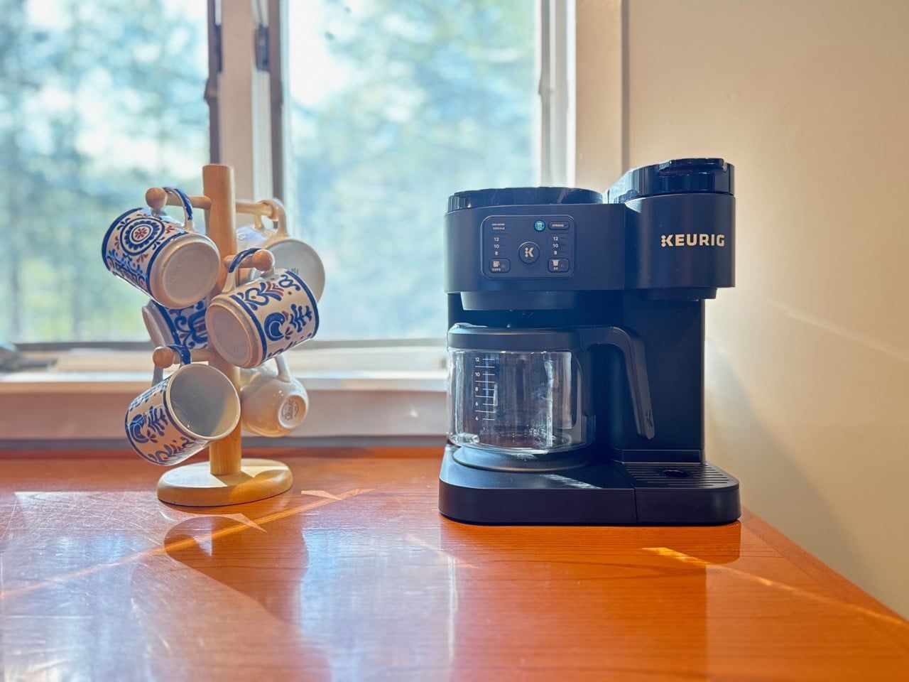 A Keurig coffee maker on a wooden table next to a mug holder with multiple mugs.