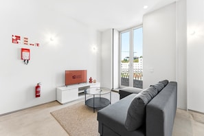 Ein modernes Wohnzimmer mit einem grauen Sofa, einem Flachbildfernseher, einem Glastisch und einem kleinen Beistelltisch. An der Wand ist ein Feuerlöscher angebracht und ein Brandschild vorhanden.