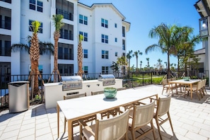 An outdoor patio with a grill, dining table, and chairs, surrounded by palm trees and a multi-story building in the background. - Landing | Amazing 2BD in East Naples (Naples)