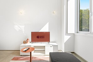 A modern hotel room with a white desk, a television displaying 'GUEST READY,' a small table with a remote control, and a window with a view of greenery. - GuestReady- Rou27- D (Porto)