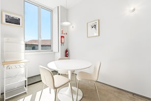 A room with a round white table, two chairs, a white shelving unit, and a window with a view of buildings. - GuestReady- Rou27- D (Porto)