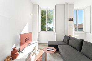 A modern living room with a grey sofa, a flat-screen TV, a small table with a remote, and a window with a view of trees and buildings. - GuestReady- Rou27- D (Porto)
