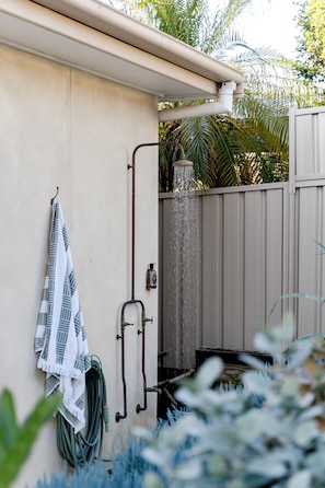 A wall-mounted shower with a rain showerhead, a towel hanging on a hook, and a garden hose coiled against the wall. - Salted Sun with pool, spa & fire pit (Manyana)