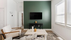 Een moderne woonkamer met een flatscreen televisie aan een groene muur, een salontafel met decoratieve objecten, en een zithoek met een witte bank en fauteuil.