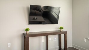 A modern living room with a flat-screen TV mounted on the wall, a wooden console table with two potted plants, and a sofa. - Landing | Stylish 2BD, Gym, Pool (Mission Bend)