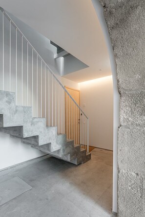 A modern staircase with a concrete finish and white railings, leading to a room with a stone wall and a door. - GuestReady- Rou2-E (Porto)