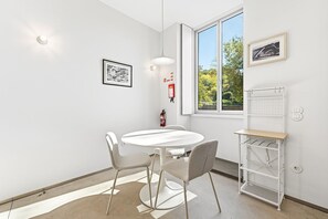 A room with a round table, two chairs, a pendant light, a window with a view of trees, a fire extinguisher, and a white shelf unit. - GuestReady- Rou2-E (Porto)