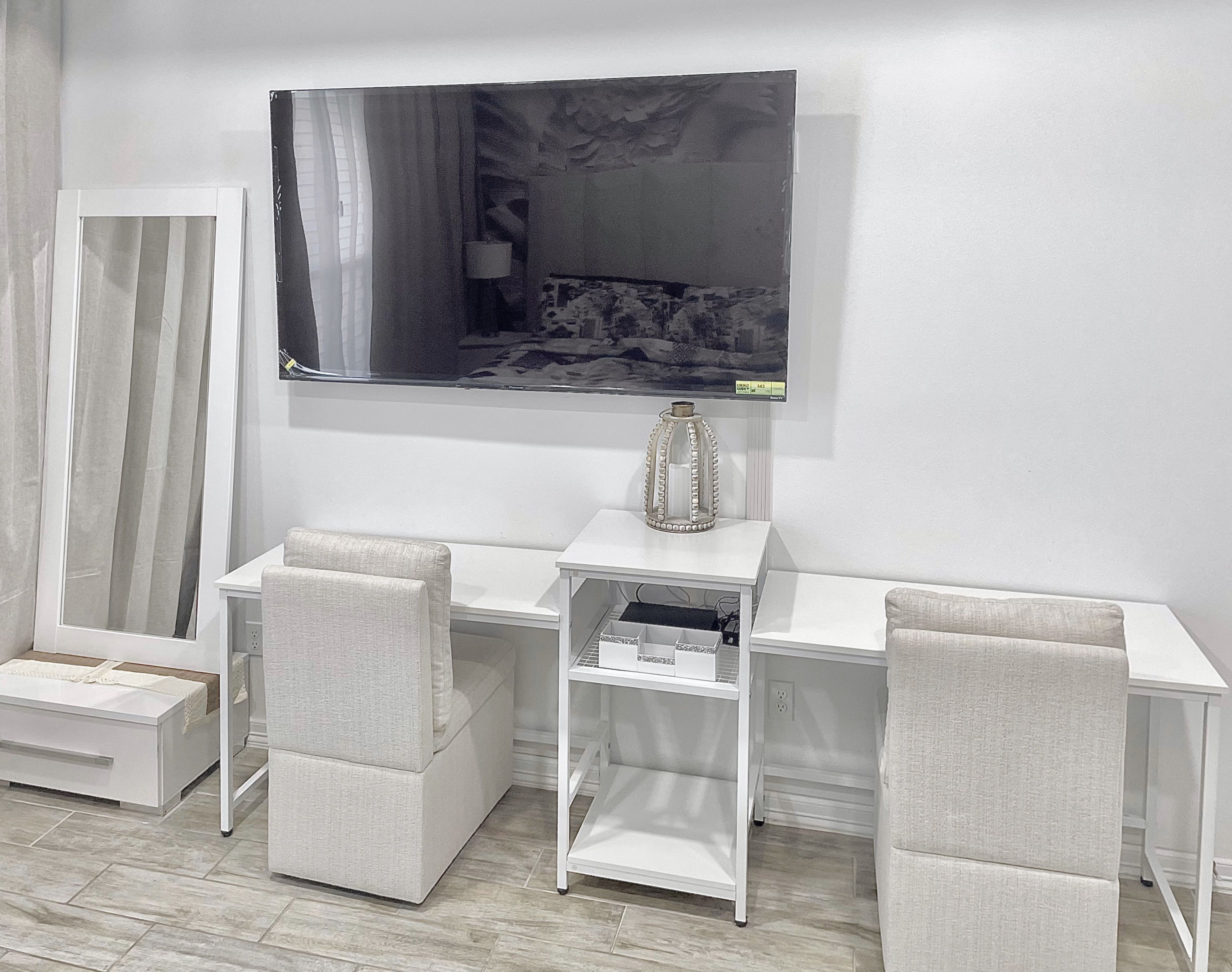 Quarto moderno com escrivaninha branca, duas cadeiras, televisão na parede e um vaso decorativo.