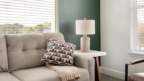 A room with a grey sofa, a patterned pillow, a lamp on a side table, and a view through window blinds. - Landing | Incredible 2BD, Pool, Gym (Mission Bend)