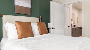 A hotel room with a bed, two pillows, a nightstand, a lamp, and a door leading to a bathroom. - Landing | Modern 2BD, Pool, Gym (Mission Bend)
