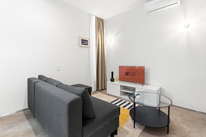 Ein modernes Wohnzimmer mit einem grauen Sofa, einem Glastisch, einem Flachbildfernseher und einem kleinen Beistelltisch.