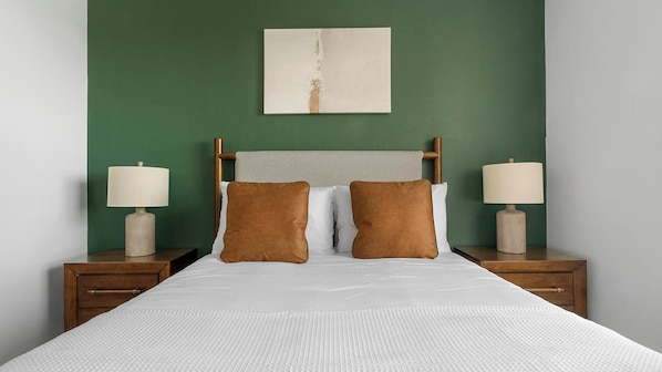 A bed with a white mattress and two brown pillows, flanked by wooden nightstands with lamps, against a green wall with a simple artwork above. - Landing | Modern 1BD in Sunnyside (Houston)