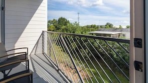 A balcony with a metal railing, a chair, and a view of a residential area with trees and a house. - Landing | Modern 1BD in Sunnyside (Houston)