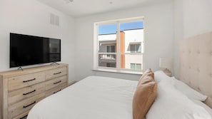 A bedroom with a bed, a TV on a wooden dresser, a window with a view of buildings, and a white wall. - Landing | Spectacular 2BD, Gym (Phoenix)
