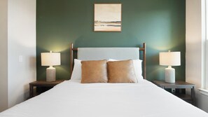 Een keurig opgemaakt bed met twee kussens, met aan weerszijden twee nachtkastjes met lampen, in een kamer met een groene muur en een schilderij boven het bed.