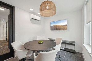A conference room with a round table, white chairs, a hanging light fixture, an air conditioning unit, and a landscape painting on the wall. - Landing | Beautiful 1BD, Clubhouse, Pool (Sarasota)
