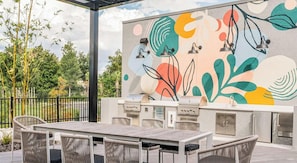 An outdoor patio with a grill, dining table, and chairs, and a colorful abstract mural on the wall. - Landing | Spectacular 1BD in North Kissimmee (Kissimmee)