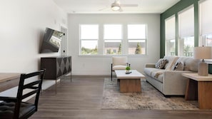 A modern living room with a sofa, coffee table, and TV, large windows, and a ceiling fan. - Landing | Amazing 2BD in North Kissimmee (Kissimmee)