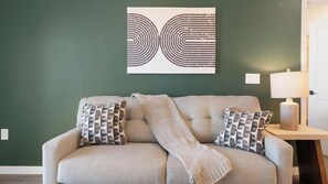 A living room with a sofa, patterned pillows, a throw blanket, a wall art piece, a lamp, and a wooden side table. - Landing | Incredible 2BD in North Kissimmee (Kissimmee)