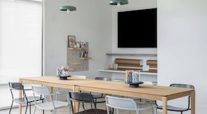 A modern meeting room with a long wooden table, six chairs, a mounted television, and wall-mounted shelves with office supplies. - Landing | Modern 2BD in North Kissimmee (Kissimmee)