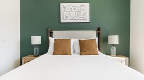 A neatly made bed with white bedding and two brown pillows, flanked by two bedside tables with lamps, against a green wall with a framed artwork above. - Landing | Spectacular 2BD in North Kissimmee (Kissimmee)