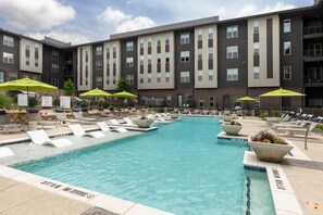 A swimming pool with lounge chairs and umbrellas, surrounded by a multi-story building with a brick facade. - Landing | Incredible 2BD, Courtyard (Garland)