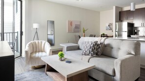 A modern living room with a sofa, a coffee table, and a chair. There is a kitchen area with cabinets and a refrigerator. - Landing | Stunning 1BD, Gym (Birmingham)